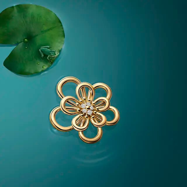 Image showcasing a delicate gold flower Van Cleef & Arpels pendant with diamond detailing, set against a serene blue backdrop with a lily pad and a tiny frog, evoking a sense of nature-inspired luxury.